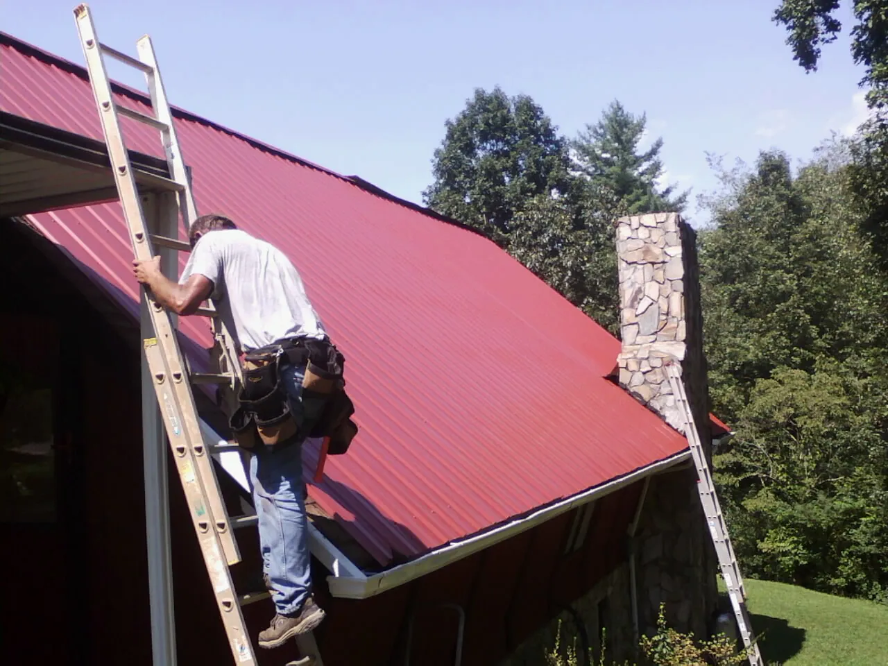 Roofer on a ladder beside a red metal roof and stone chimney — Soffit & Fascia Repair by Roofing Brothers in Orbisonia